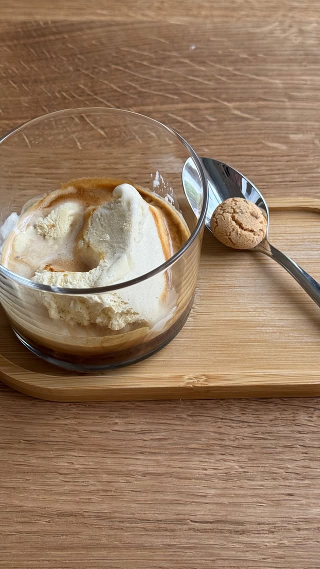 Ein Affogato: eine Kugel Vanilleeis mit Espresso übergossen in einem Glas, daneben ein Löffel mit einem Amaretto-Keks auf einem Holzbrett.