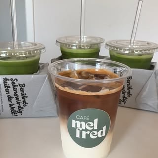 Vier Becher to go: ein Iced Coffee mit dem grünen „mel fred“-Logo vorn, dahinter drei Becher mit grünem Iced Matcha.