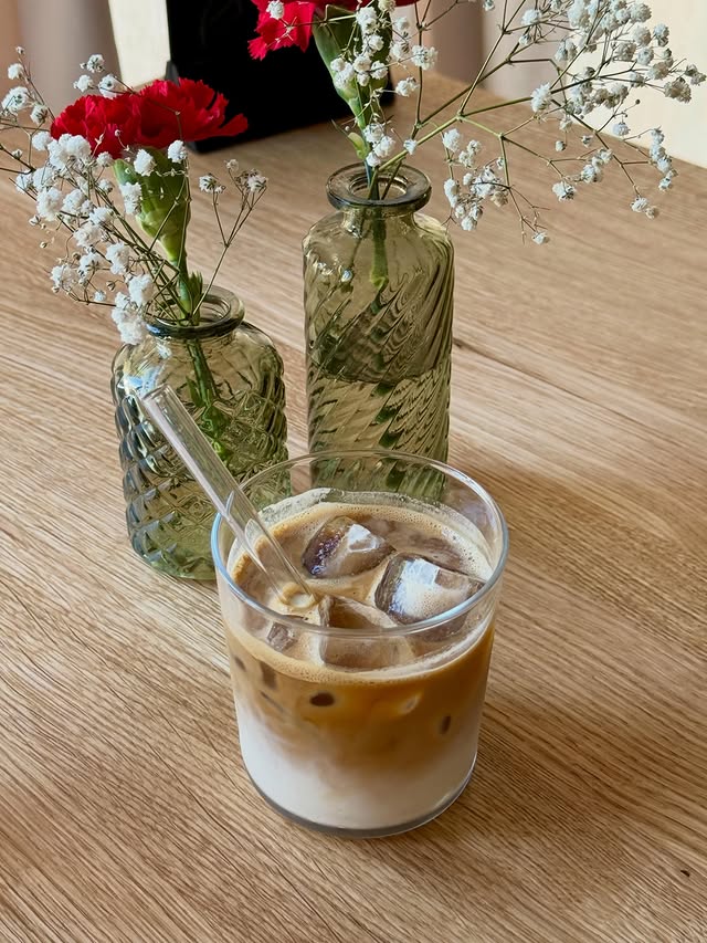 Ein Iced Coffee mit Milch im Glas mit Glas-Trinkhalm auf einem hellen Holztisch, daneben zwei grüne Vasen mit Nelken und Schleierkraut.