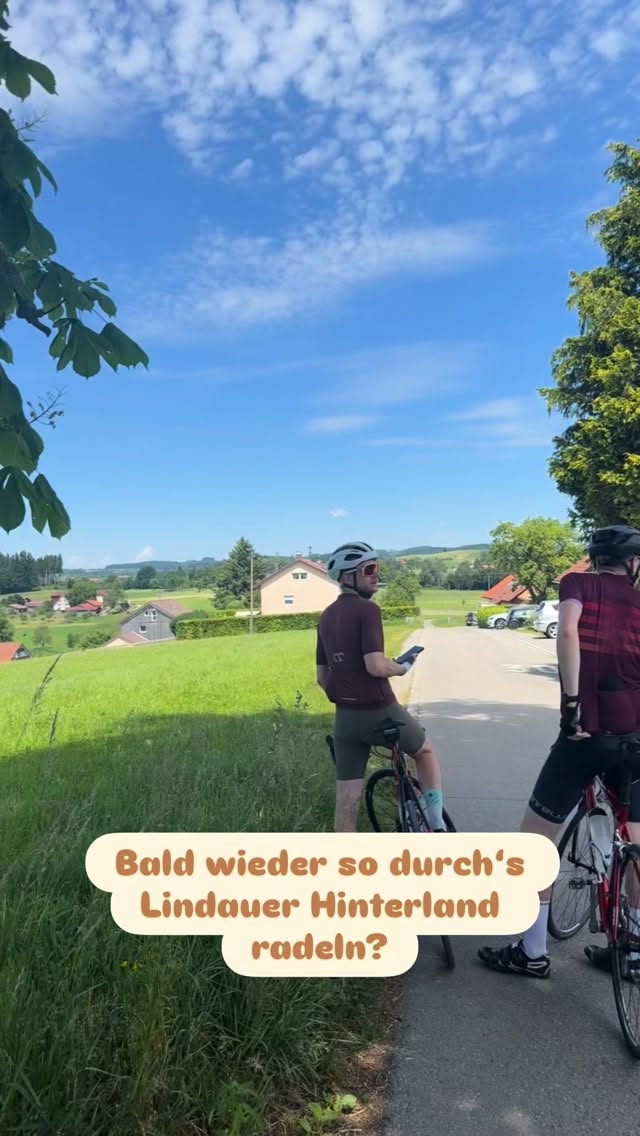 Blick über das grüne Lindauer Hinterland: Wiesen, vereinzelte Häuser und Hügel unter einem blauen Sommerhimmel, im Vordergrund zwei Rennradfahrer auf einer kleinen Straße.