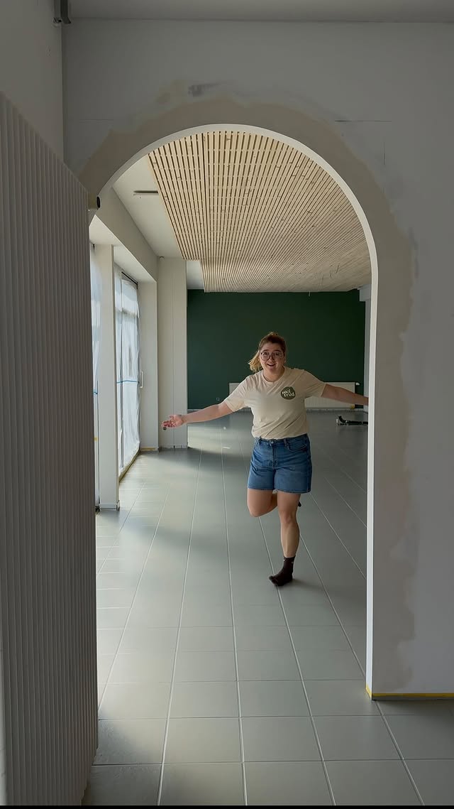 Blick durch einen weißen Rundbogen in den hellen Café-Raum mit Holz-Lamellendecke und grüner Wand; eine Person im „mel fred“-Shirt tanzt fröhlich hindurch.