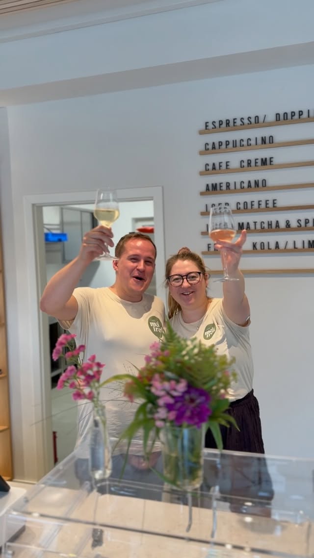 Zwei Gründer:innen in cremefarbenen „mel fred“-Shirts stoßen lachend mit Sektgläsern an; an der Wand hinter ihnen die Kaffeekarte mit Espresso, Cappuccino, Café Crème, Americano, Iced Coffee und Matcha.